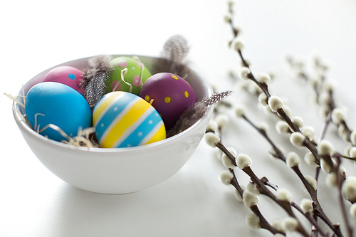holidays and object concept - colored easter eggs with quail feathers in bowl and pussy willow branches on white background