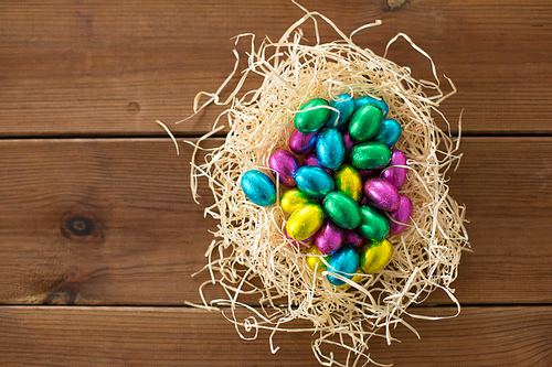 easter, confectionery and holidays concept - chocolate eggs in foil wrappers in straw nest on wooden background