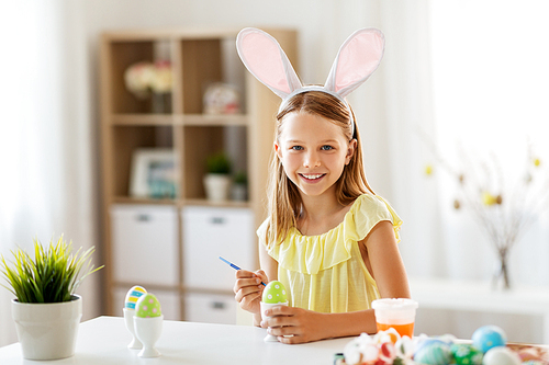 easter, holidays and people concept - happy girl wearing bunny ears headband coloring eggs with colors and brush at home