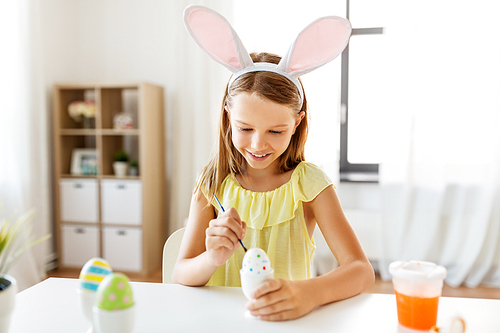 easter, holidays and people concept - happy girl wearing bunny ears headband coloring eggs with colors and brush at home