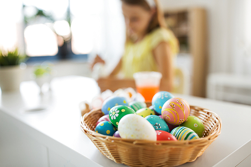 easter, holidays and people concept - colored eggs in wicker basket and girl on background at home