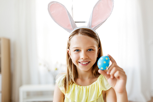 easter, holidays and people concept - happy girl wearing bunny ears headband with colored egg at home