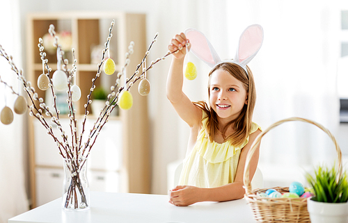 easter, holidays and people concept - happy girl wearing bunny ears headband decorating willow branches by toy eggs at home