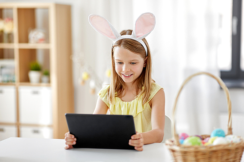 easter, holidays and people concept - happy girl wearing bunny ears headband with tablet pc computer and basket of colored eggs at home