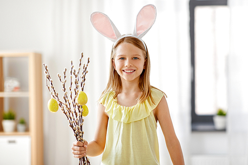 easter, holidays and people concept - happy girl wearing bunny ears headband holding willow branches decorated toy eggs at home