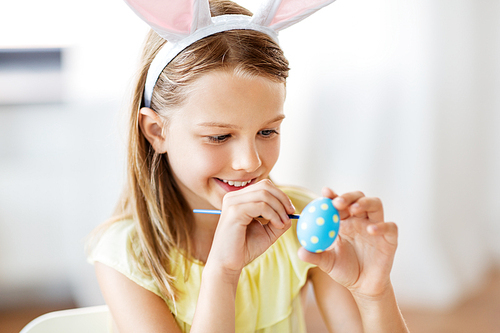 easter, holidays and people concept - happy girl wearing bunny ears headband coloring egg by brush at home