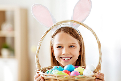 easter, holidays and people concept - happy girl wearing bunny ears headband with basket of colored eggs at home
