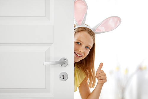 easter, holidays and people concept - happy girl wearing bunny ears peeking out door and showing thumbs up at home