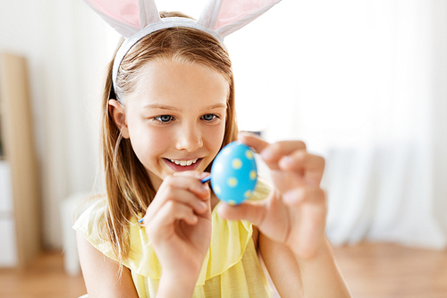 easter, holidays and people concept - happy girl wearing bunny ears headband coloring egg by brush at home