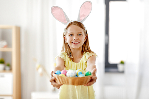 easter, holidays and people concept - happy girl wearing bunny ears headband with basket of colored eggs at home