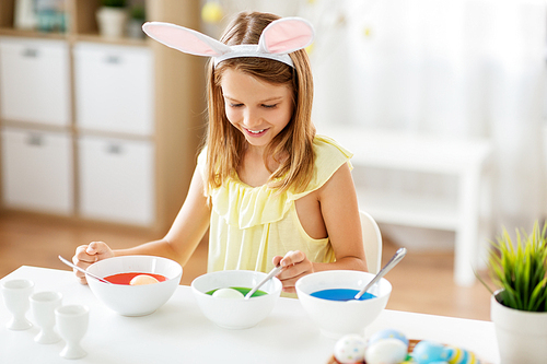 easter, holidays and people concept - happy girl wearing bunny ears headband coloring eggs by liquid dye at home