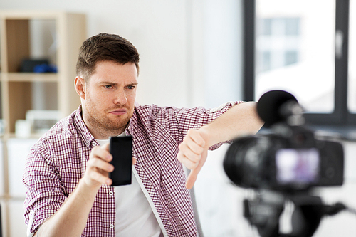 blogging, videoblog and people concept - displeased male . with camera recording video review of smartphone and showing thumbs down gesture at home office