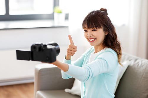 blogging, videoblog and technology concept - asian woman or . with camera recording video blog or taking selfie and showing thumbs up at home