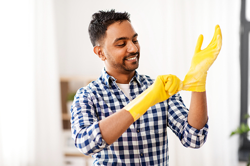 cleaning, housework and housekeeping concept - smiling indian man putting protective rubber gloves on at home