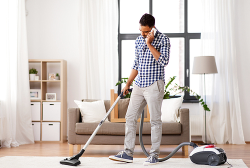 household and cleaning concept - indian man with vacuum cleaner calling on smartphone at home