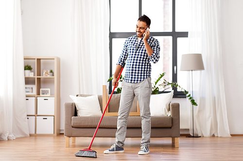 cleaning, housework and housekeeping concept - indian man with broom sweeping floor and calling on smartphone at home