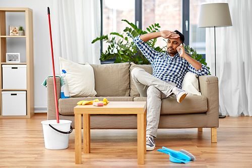 household and technology concept - tired indian man calling on smartphone after cleaning home