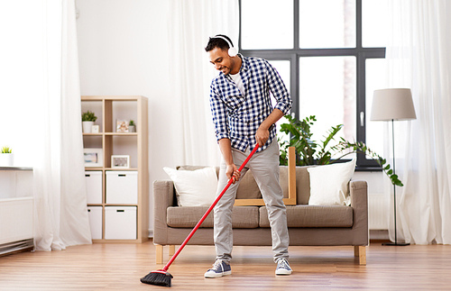 cleaning, housework and housekeeping concept - indian man in headphones with broom sweeping floor at home