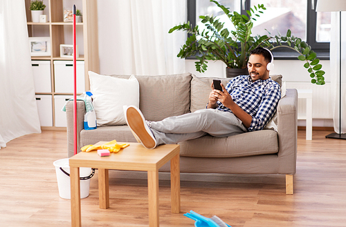 people, housework and housekeeping concept - indian man in headphones listening to music on smartphone after home cleaning