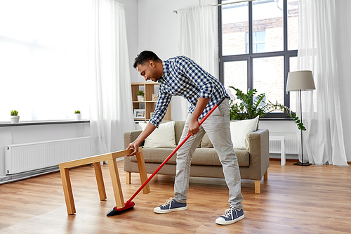 cleaning, housework and housekeeping concept - indian man with broom sweeping floor under table at home