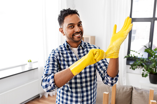 cleaning, housework and housekeeping concept - smiling indian man putting protective rubber gloves on at home