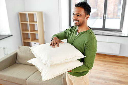 household, moving and cleaning concept - happy indian man holding two pillows or sofa cushion at home