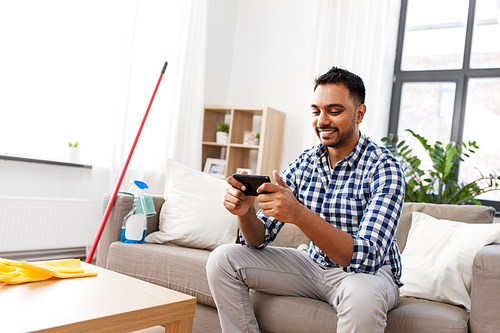 household and technology concept - smiling indian man playing game on smartphone after cleaning home