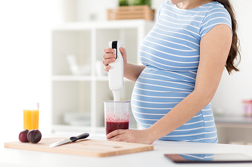 cooking, pregnancy and healthy eating concept - pregnant woman with blender making fruit smoothie drink at home kitchen