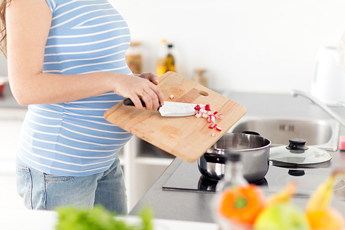 pregnancy, cooking food and healthy eating concept - close up of pregnant woman with kitchen knife adding chopped raddish ftom wooden cutting board to pot on stove at home