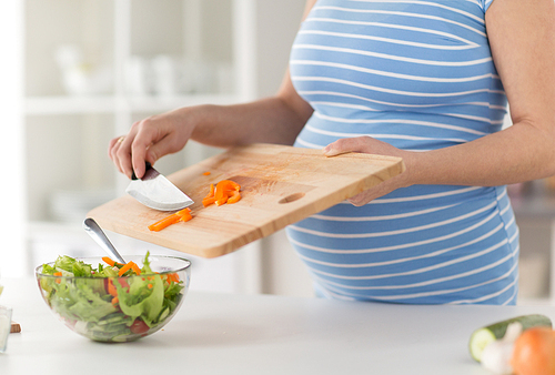 pregnancy, cooking food and healthy eating concept - close up of pregnant woman with kitchen knife adding chopped pepper from wooden cutting board to salad bowl at home