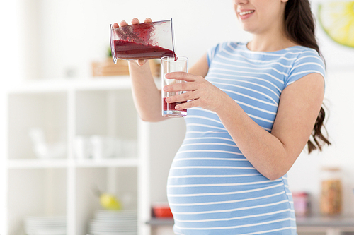 healthy eating, cooking and pregnancy concept - pregnant woman pouring fruit smoothie from blender cup to glass and at home kitchen