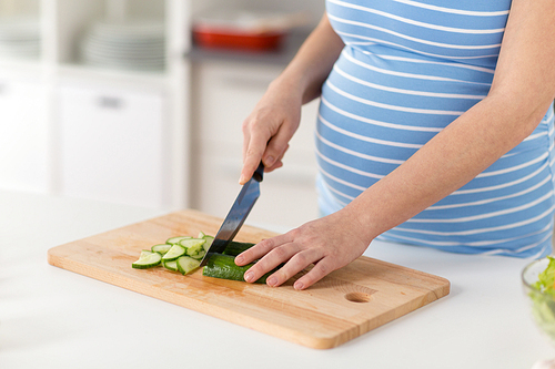 pregnancy, cooking food and healthy eating concept - close up of pregnant woman chopping cucumber by kitchen knife on wooden cutting board at home