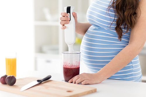 cooking, pregnancy and healthy eating concept - pregnant woman with blender making fruit smoothie drink at home kitchen