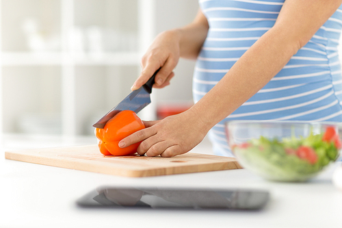 pregnancy, cooking food and healthy eating concept - close up of pregnant woman making vegetable salad and chopping pepper by kitchen knife on cutting board at home kitchen