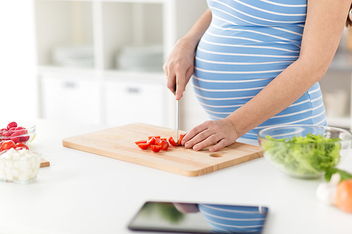 pregnancy, cooking food and healthy eating concept - close up of pregnant woman with kitchen knife chopping tomato on wooden cutting board at home