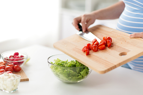 pregnancy, cooking food and healthy eating concept - close up of pregnant woman with kitchen knife adding chopped cherry tomato from wooden cutting board to salad bowl at home