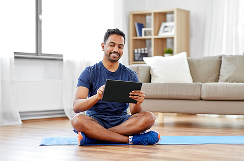 sport, technology and healthy lifestyle concept - smiling indian man with tablet computer sitting on exercise mat at home