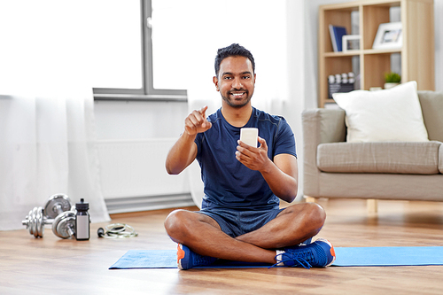 sport, technology and healthy lifestyle concept - smiling indian man with smartphone sitting on exercise mat and pointing to you at home