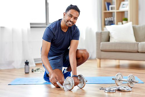 fitness, sport, weightlifting and bodybuilding concept - smiling indian man assembling dumbbells at home