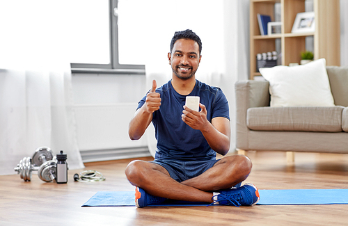 sport, technology and healthy lifestyle concept - smiling indian man with smartphone sitting on exercise mat and showing thumbs up at home