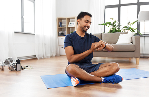 sport, technology and healthy lifestyle concept - smiling indian man with fitness tracker sitting on exercise mat at home