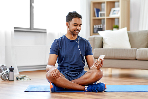 sport, technology and healthy lifestyle concept - smiling indian man in earphones listening to music on smartphone at home