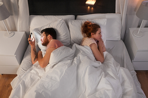 technology, internet addiction and cheat concept - man using smartphone at night while his girlfriend is sleeping