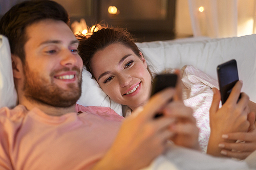 technology, internet and communication concept - happy couple using smartphones in bed at night