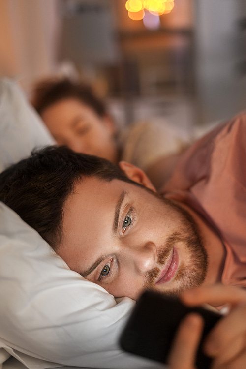 technology, internet addiction and cheat concept - man using smartphone at night while his girlfriend is sleeping