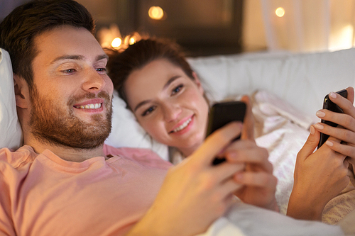 technology, internet and communication concept - happy couple using smartphones in bed at night