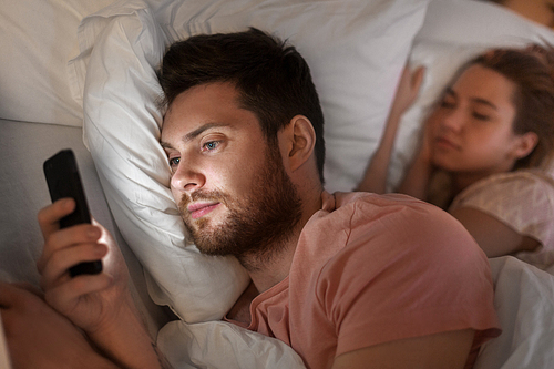 technology, internet addiction and cheat concept - man using smartphone at night while his girlfriend is sleeping