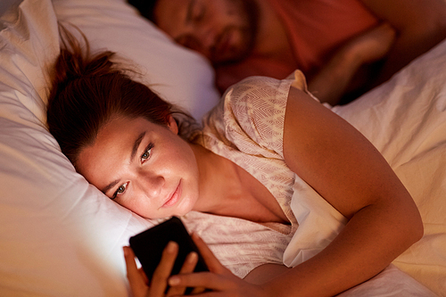 technology, internet addiction and cheat concept - woman using smartphone at night while her boyfriend is sleeping