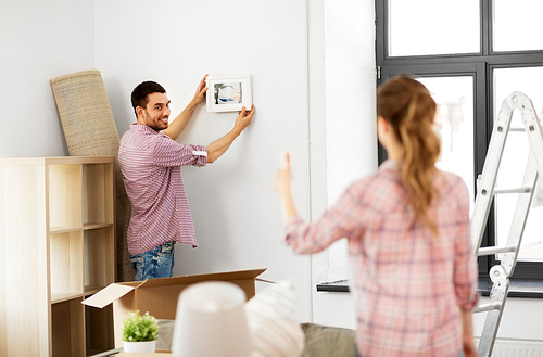 repair, accommodation and real estate concept - smiling couple moving to new home and hanging photo on wall