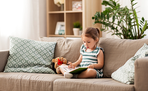 childhood and people concept - little girl sitting on sofa reading book and toy teddy bear at home